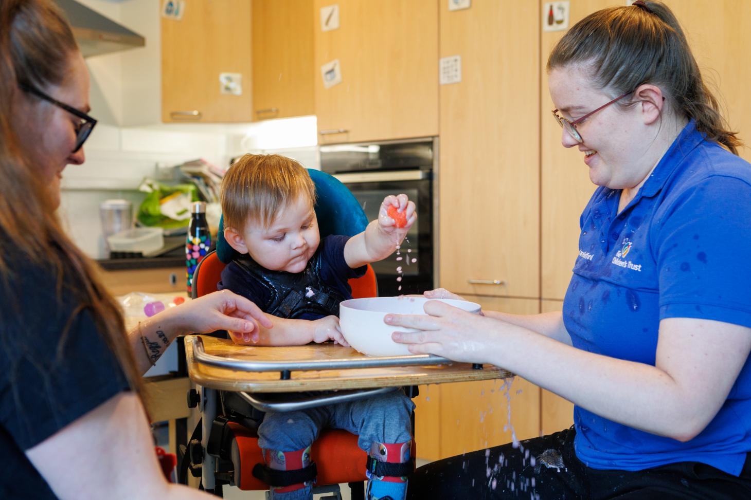 A young boy in a supportive chair reaches toward a bowl held by a therapist during an activity.