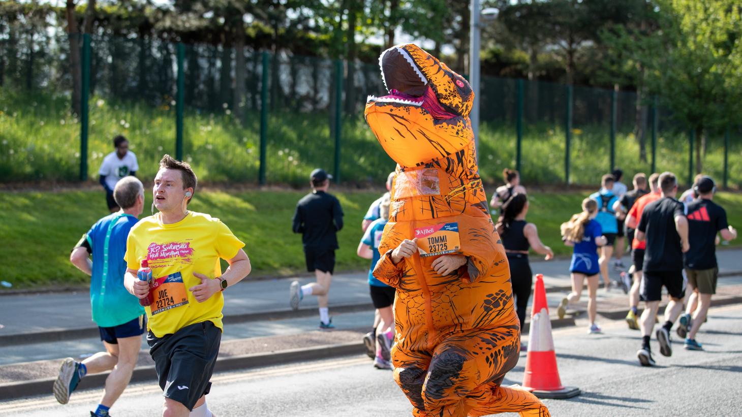 runners and a runner in an inflatable orange dinosaur costume running on a road in front of a grass bank and green fencing