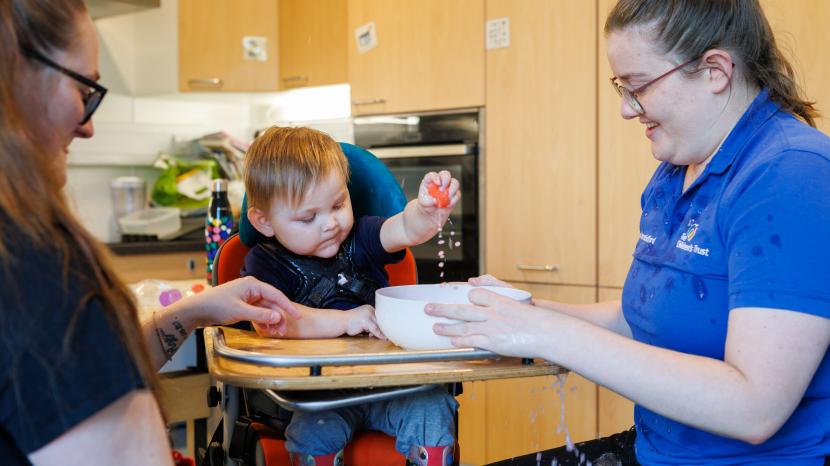 A young boy in a supportive chair reaches toward a bowl held by a therapist during an activity.